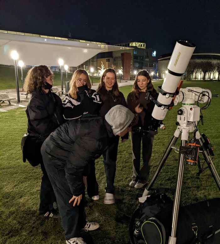 Foto van een groep vrolijk kijkende jongeren, waarvan één door de telescoop aan het kijken is.