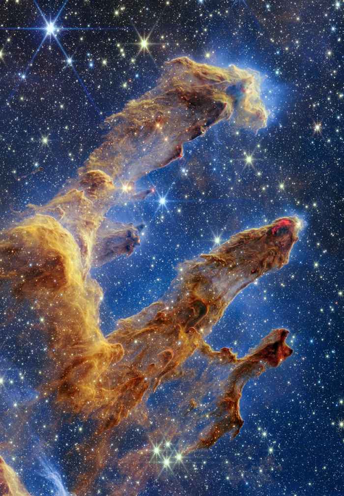 The pillars of creation, a mustard coloured nebula, against a blue background. The nebula two quite distinct an one slightly less pronounced pillars of material.
