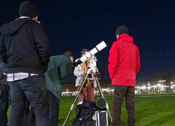A telescope surrounded by people, one of whom (a young man) is looking through the telescope