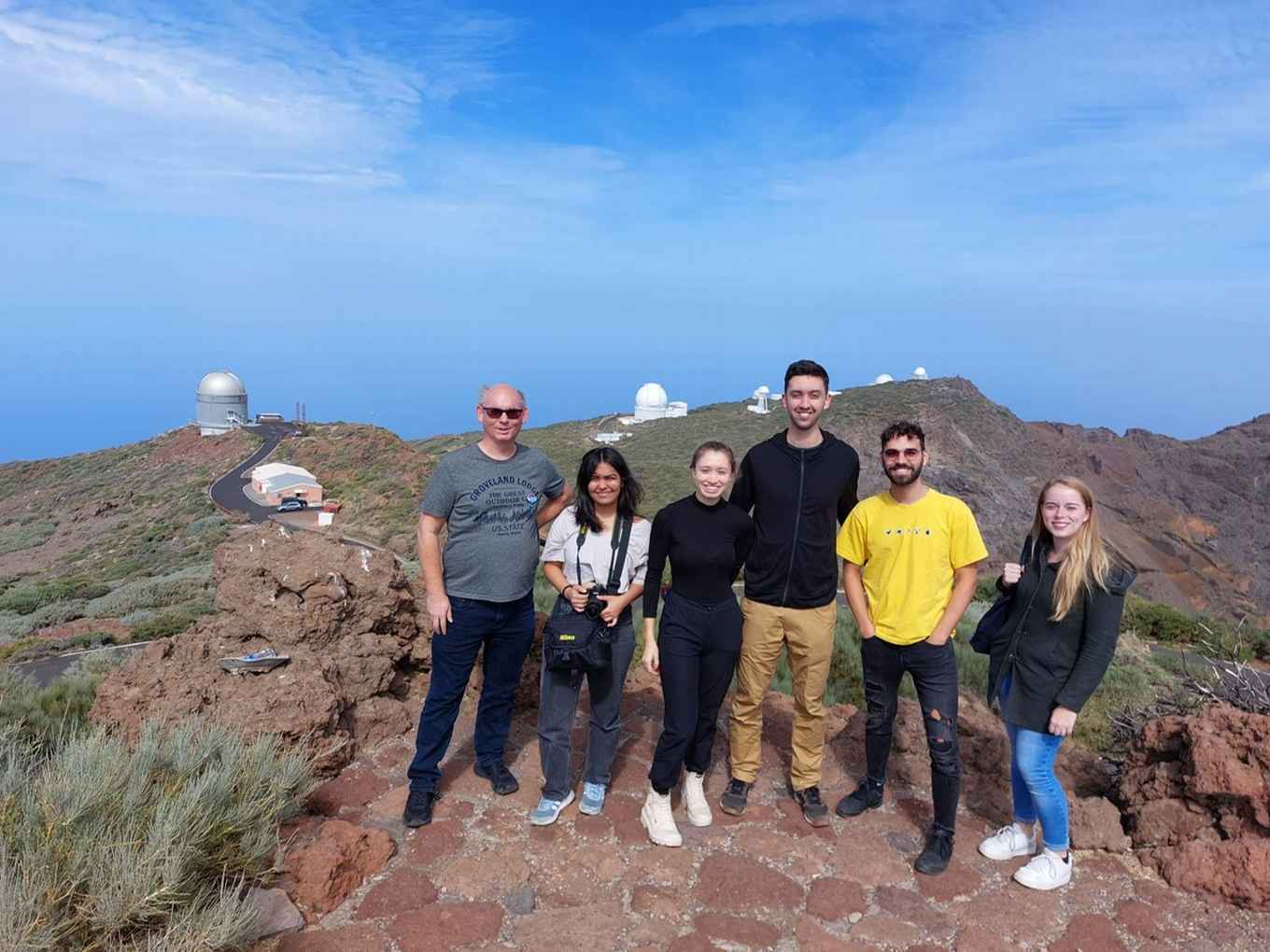 Een docent met een groep studenten op een berg met blauwe hemel op de achtergrond. Op een berg verderop staan telescoopkoepels.